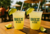 Two Breeze & Co. cold drink cups with lime wedges on a wooden outdoor table under umbrellas.