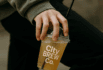 A person holding a City Brew Co. iced drink cup with a black straw in an urban outdoor setting.