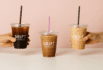 Three iced coffee cups from Drift Coffee Co., each held by a hand against a soft pink background.