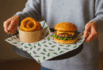 Premium custom food liner with green chili pattern under cheeseburger and onion rings on a black tray held by a person.