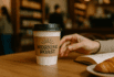 Coffee cup with Morning Roast kraft sleeve, hand reaching on wooden café table.