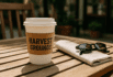 Coffee cup with Harvest Grounds sleeve placed beside sunglasses and newspaper on a wooden patio table.