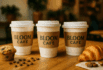 Three coffee cups with Bloom Café sleeves on a wooden table, surrounded by coffee beans and pastries.