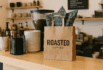 Brown kraft paper bag with Roasted Coffee logo on a coffee shop counter surrounded by plates and coffee equipment.