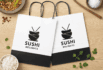 Two white paper takeout bags with Sushi Delights logo, placed on a wood surface with bowls of rice and herbs.