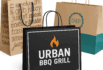Three branded takeout bags including Urban BBQ Grill’s black bag with flame logo and kraft bags with grill-themed prints.
